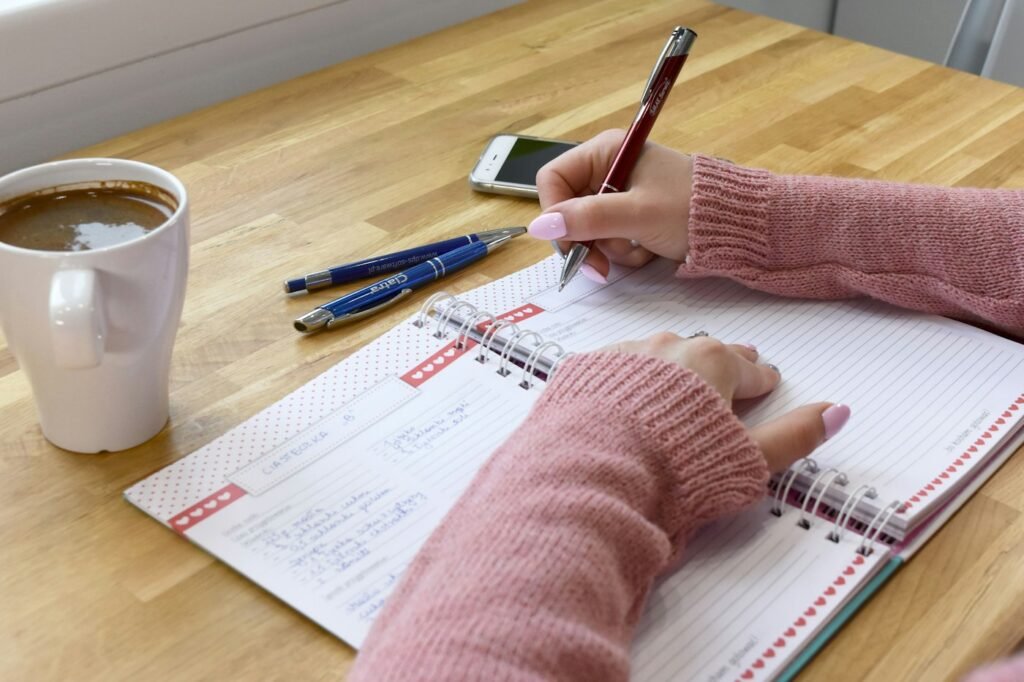 Person writing a weekly plan in a notebook beside a cup of coffee