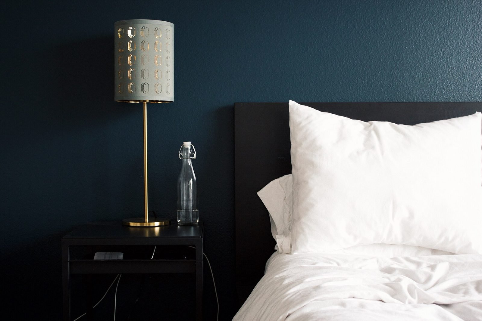 A calm bedroom with a bedside lamp, a glass water bottle on the nightstand, and white pillows against a dark blue wall